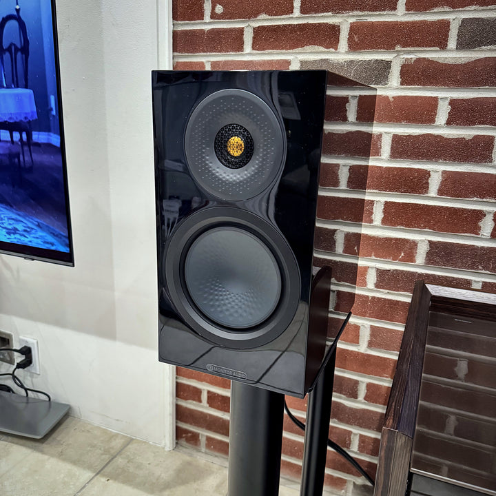 Monitor Audio Silver 8G two-way bookshelf speaker in gloss black on a black stand, showing its gold-dome tweeter and dimpled bass driver