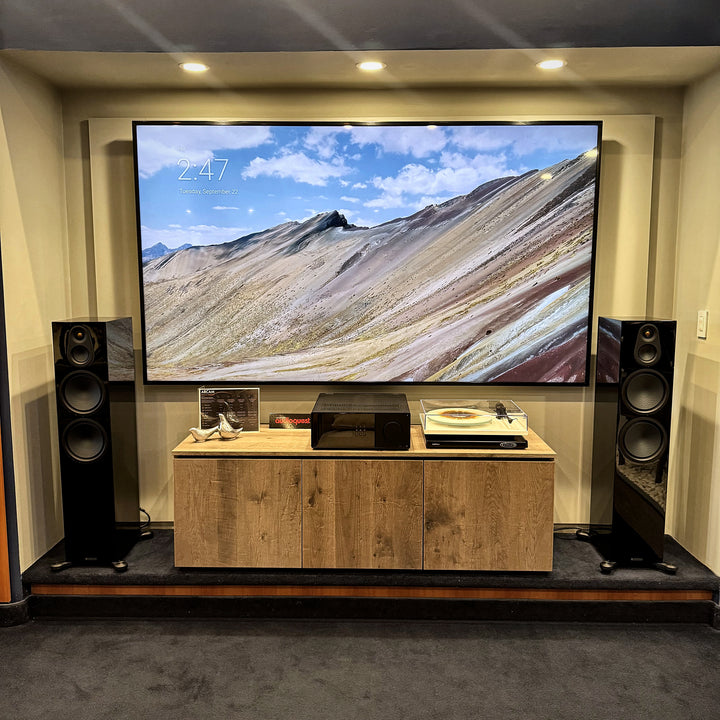 Pair of Monitor Audio Silver 8G floorstanding speakers in gloss black on either side of a large TV and an oak media cabinet